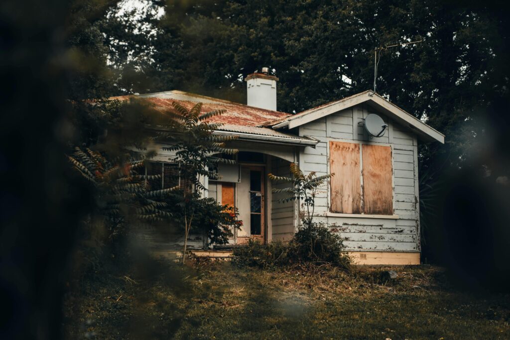 Vacant boarded house with overgrown yard showing signs of neglect and deterioration from prolonged holding time in real estate investment.