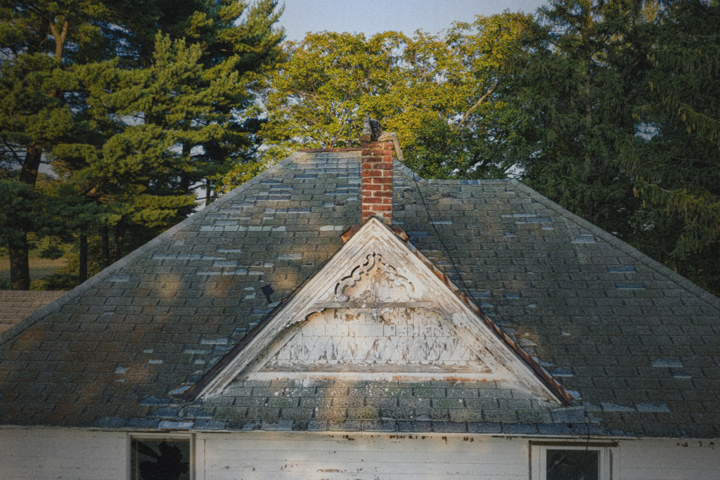 A weathered roof on a distressed property showing aging shingles, deteriorating trim, and signs of deferred maintenance surrounded by trees.