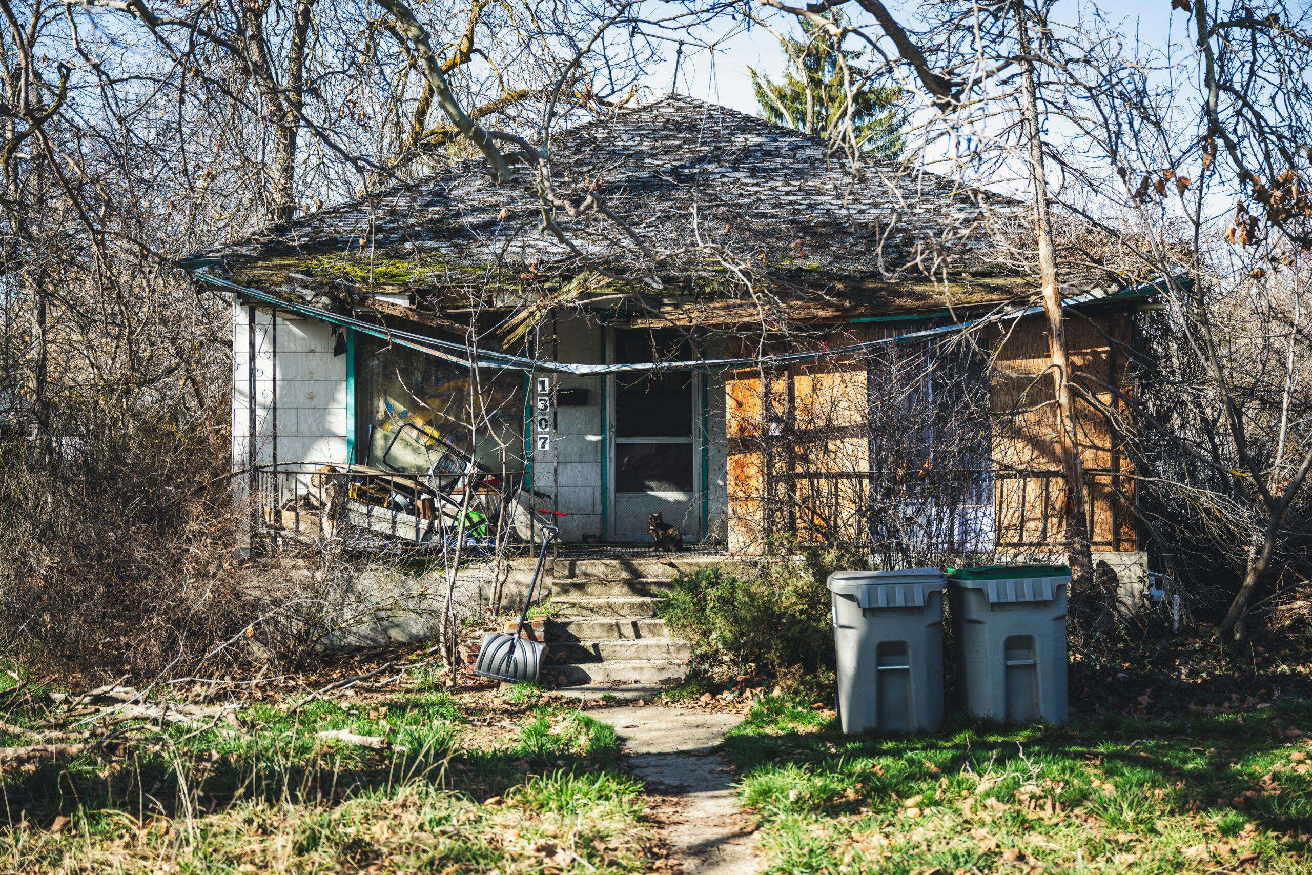 Vacant distressed house with roof deterioration, overgrown vegetation, boarded windows, and visible signs of neglect caused by a lack of property preservation.